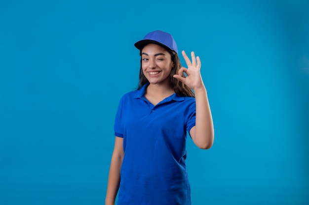 Giovane ragazza di consegna in uniforme blu e cappuccio che guarda l'obbiettivo ammiccante sorridendo allegramente facendo segno ok in piedi su sfondo blu