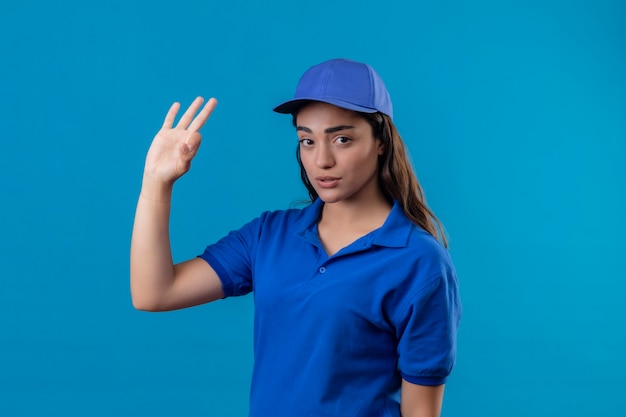 Giovane ragazza di consegna in uniforme blu e cappuccio che guarda l'obbiettivo con l'espressione triste sul viso facendo segno ok in piedi su sfondo blu
