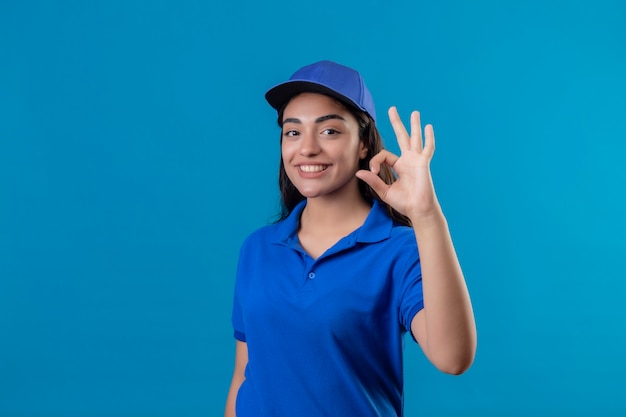 Giovane ragazza di consegna in uniforme blu e cappuccio che guarda l'obbiettivo sorridente allegramente facendo segno giusto in piedi su sfondo blu