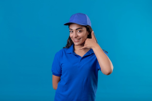 Giovane ragazza di consegna in uniforme blu e cappuccio che guarda l'obbiettivo sorridente fiducioso che mi chiama gesto in piedi su sfondo blu