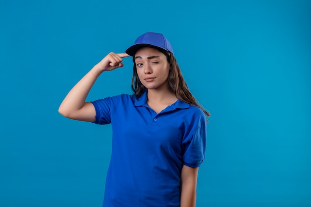 Giovane ragazza di consegna in uniforme blu e cappuccio che indica tempio ammiccante che guarda l'obbiettivo con espressione fiduciosa focalizzata sul compito in piedi su sfondo blu