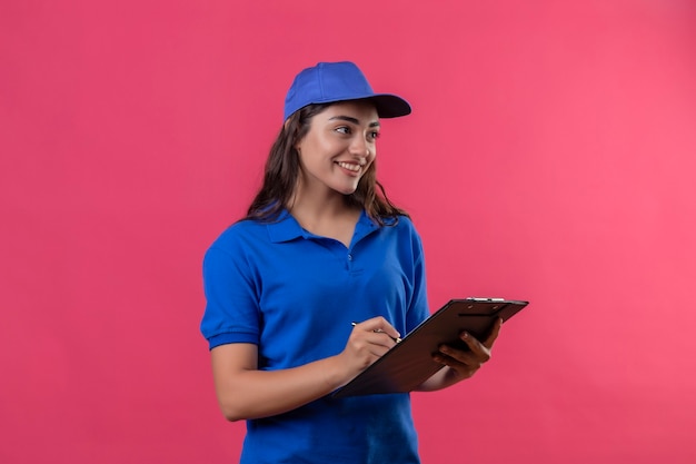 Giovane ragazza di consegna in uniforme blu e cappuccio che tiene appunti con la penna che osserva da parte sorridente fiducioso in piedi su sfondo rosa