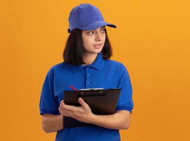 Giovane ragazza di consegna in uniforme blu e cappuccio che tiene appunti con pagine vuote e matita che guarda da parte con la faccia seria che sta sopra la parete arancione