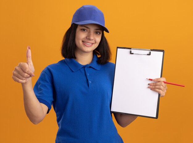 Giovane ragazza di consegna in uniforme blu e cappuccio che tiene appunti con pagine vuote e matita sorridente che mostra i pollici in su in piedi sopra la parete arancione