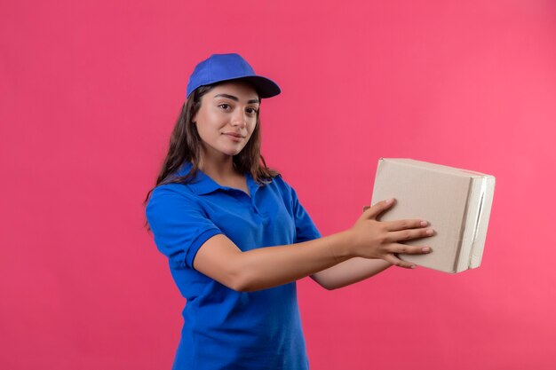 Giovane ragazza di consegna in uniforme blu e cappuccio che tiene dando il pacchetto della scatola a un cliente sorridente fiducioso in piedi su sfondo rosa