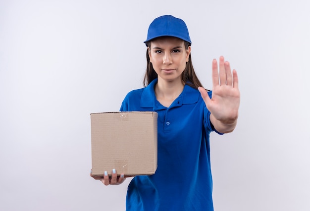 Giovane ragazza di consegna in uniforme blu e cappuccio che tiene il pacchetto della scatola che fa il segnale di stop con la mano che guarda l'obbiettivo con la faccia seria