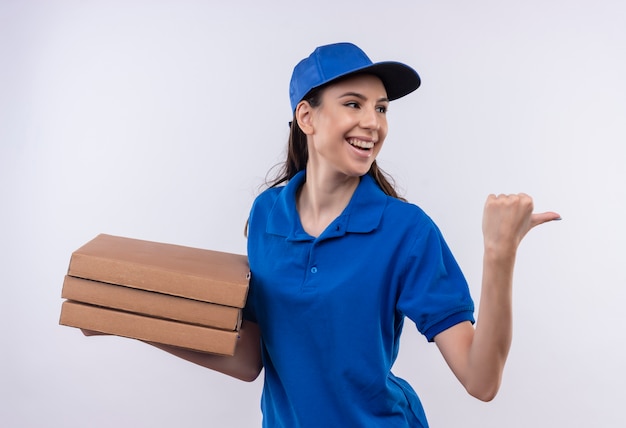 Giovane ragazza di consegna in uniforme blu e cappuccio che tiene la pila di scatole per pizza che punta indietro con il pollice che sorride allegramente