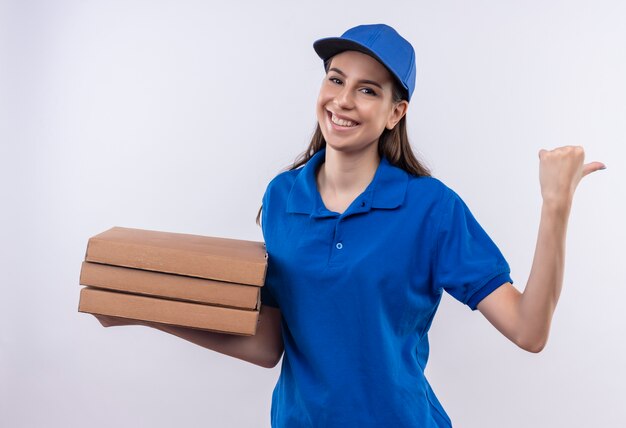 Giovane ragazza di consegna in uniforme blu e cappuccio che tiene la pila di scatole per pizza che punta indietro con il pollice che sorride allegramente