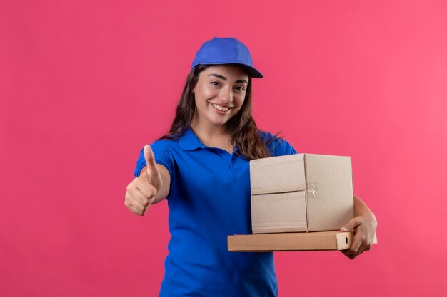 Giovane ragazza di consegna in uniforme blu e cappuccio che tiene le scatole di cartone sorridendo allegramente mostrando i pollici in su in piedi su sfondo rosa