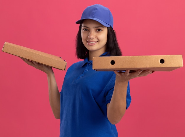 Giovane ragazza di consegna in uniforme blu e cappuccio che tiene le scatole per pizza con la faccia felice sorridente amichevole in piedi sopra la parete rosa