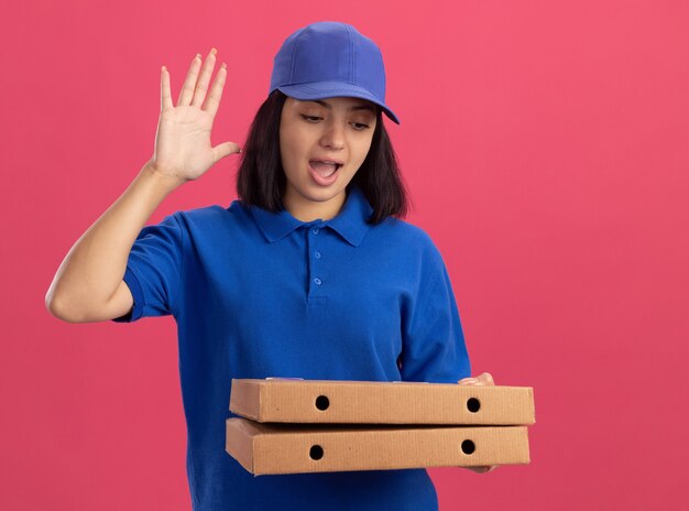 Giovane ragazza di consegna in uniforme blu e cappuccio che tiene le scatole per pizza felice e positivo che solleva la palma che sta sopra la parete rosa
