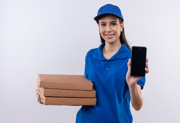 Giovane ragazza di consegna in uniforme blu e cappuccio che tiene pila di scatole per pizza che mostra smartphone sorridente fiducioso
