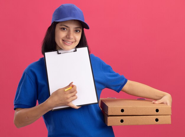 Giovane ragazza di consegna in uniforme blu e cappuccio che tiene scatole per pizza e appunti con pagine vuote in attesa di firma in piedi sopra il muro rosa