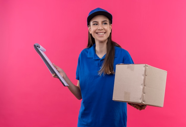 Giovane ragazza di consegna in uniforme blu e pacchetto della scatola della tenuta del cappuccio e appunti che osserva da parte con il sorriso sul fronte