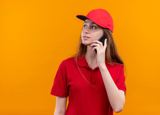 Giovane ragazza di consegna in uniforme rossa che parla sul telefono che esamina il lato sinistro sulla parete arancione isolata con lo spazio della copia