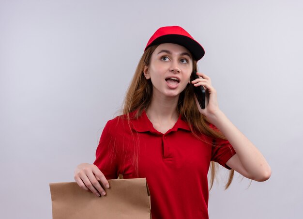 Giovane ragazza di consegna in uniforme rossa che tiene il sacchetto di carta e parla al telefono sulla parete bianca isolata