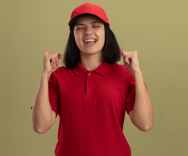 Giovane ragazza di consegna in uniforme rossa e berretto pugni di serraggio felici ed eccitati in piedi sopra la parete chiara