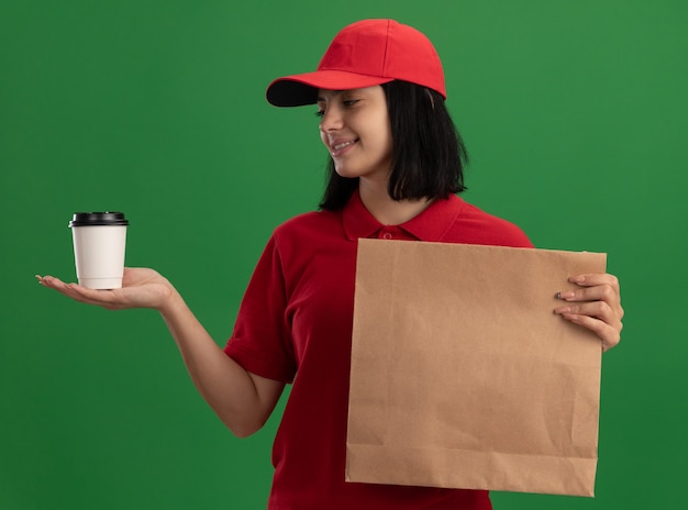 Giovane ragazza di consegna in uniforme rossa e cappuccio che tiene il pacchetto di carta e la tazza guardando la tazza che sorride con la faccia felice che sta sopra la parete verde