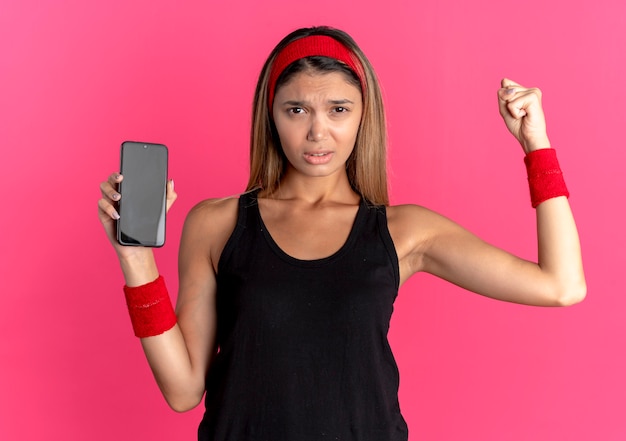 Giovane ragazza di forma fisica in abbigliamento sportivo nero e fascia rossa che mostra il pugno di serraggio dello smartphone con la faccia arrabbiata in piedi sopra la parete rosa