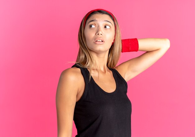 Giovane ragazza di forma fisica in abbigliamento sportivo nero e fascia rossa che osserva da parte confuso in piedi sopra la parete rosa