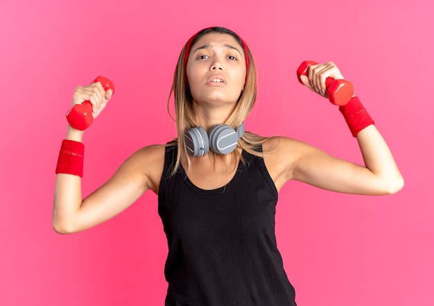 Giovane ragazza di forma fisica in abbigliamento sportivo nero e fascia rossa che risolve con i dumbbells che sembrano fiduciosi sopra il rosa