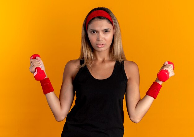Giovane ragazza di forma fisica in abbigliamento sportivo nero e fascia rossa che risolve con i dumbbells con la faccia seria che sta sopra la parete arancione