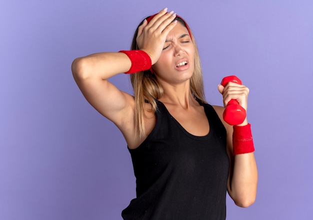 Giovane ragazza di forma fisica in abbigliamento sportivo nero e fascia rossa che tiene il manubrio che sembra stanco ed esausto con hnad sulla testa sopra il blu