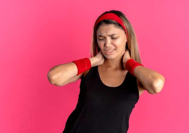 Giovane ragazza di forma fisica in abbigliamento sportivo nero e fascia rossa che tocca il suo collo che sembra malessere sensazione di dolore sul rosa