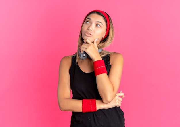 Giovane ragazza di forma fisica in abbigliamento sportivo nero e fascia rossa con le cuffie che osserva da parte con la mano sul mento perplessa sul rosa
