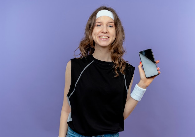 Giovane ragazza di forma fisica in abiti sportivi neri con archetto che mostra smartphone sorridente allegramente in piedi sopra la parete blu