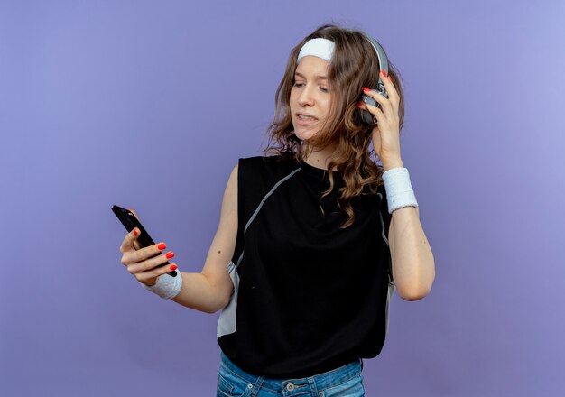 Giovane ragazza di forma fisica in abiti sportivi neri con archetto e cuffie guardando il suo smartphone alla ricerca di musica in piedi sopra la parete blu