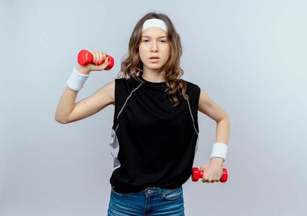 Giovane ragazza di forma fisica in abiti sportivi neri con fascia che risolve con i dumbbells che sembrano fiduciosi in piedi sopra il muro bianco