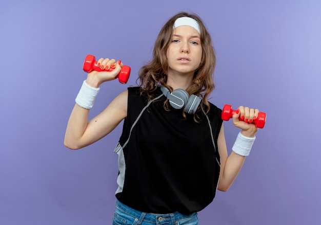 Giovane ragazza di forma fisica in abiti sportivi neri con fascia che risolve con i dumbbells che sembrano sicuri sopra l'azzurro