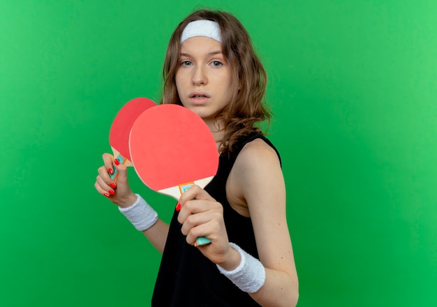 Giovane ragazza di forma fisica in abiti sportivi neri con fascia che tiene due racchette per ping-pong con faccia seria in piedi sopra la parete verde