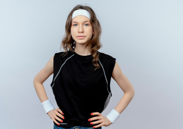 Giovane ragazza di forma fisica in abiti sportivi neri con fascia con espressione seria e sicura in piedi sopra il muro bianco