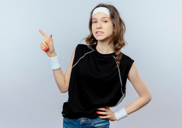 Giovane ragazza di forma fisica in abiti sportivi neri con fascia confusa puntando il dito contro qualcosa in piedi sopra il muro bianco