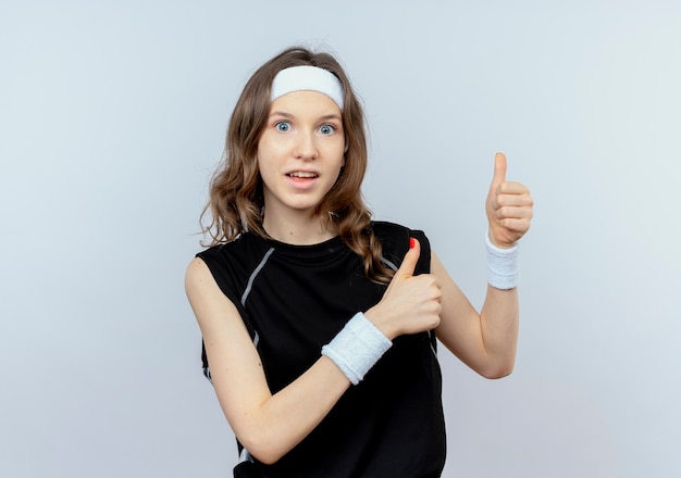 Giovane ragazza di forma fisica in abiti sportivi neri con fascia sorridente che mostra i pollici in su in piedi sul muro bianco