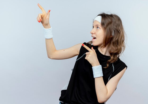 Giovane ragazza di forma fisica in abiti sportivi neri con fascia sorridente che punta con le dita a lato in piedi sul muro bianco