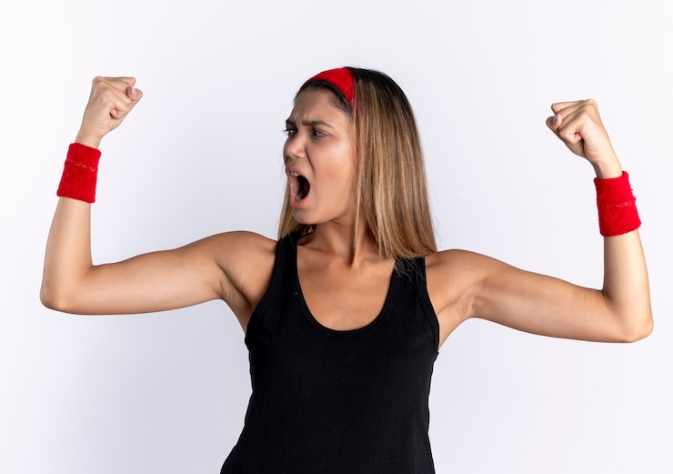 Foto gratuito giovane ragazza di forma fisica in abiti sportivi neri e fascia rossa che alza i pugni che grida con la faccia seria che grida in piedi sopra il muro bianco