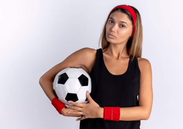 Giovane ragazza di forma fisica in abiti sportivi neri e fascia rossa che tiene pallone da calcio con la faccia seria che sta sopra il muro bianco