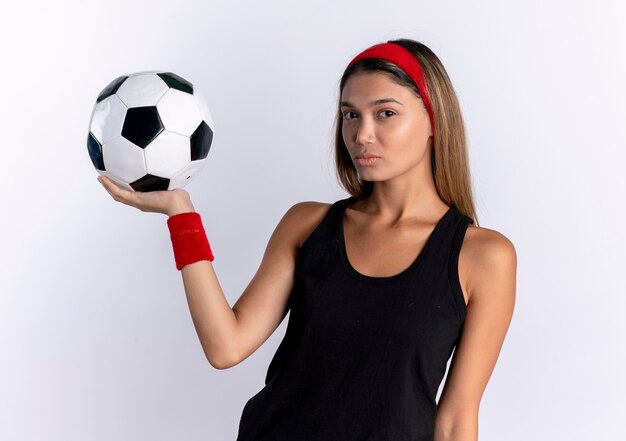 Giovane ragazza di forma fisica in abiti sportivi neri e fascia rossa che tiene pallone da calcio con la faccia seria che sta sopra il muro bianco