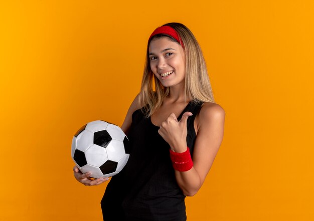 Giovane ragazza di forma fisica in abiti sportivi neri e fascia rossa che tiene pallone da calcio sorridente che mostra i pollici in su in piedi sopra la parete arancione