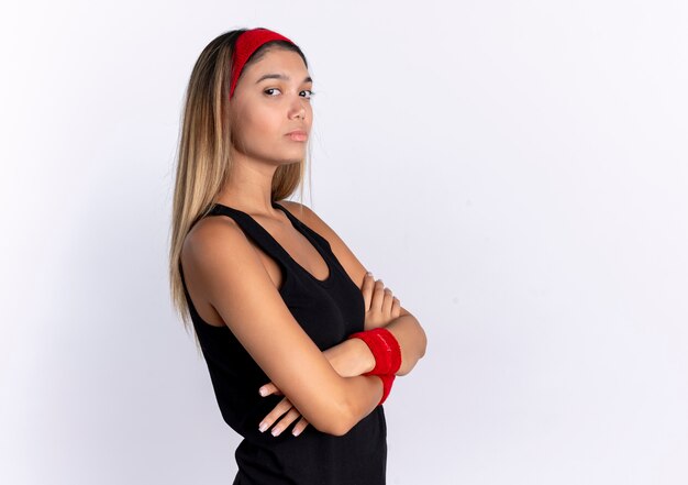 Giovane ragazza di forma fisica in abiti sportivi neri e fascia rossa con espressione seria e sicura con le braccia incrociate in piedi sul muro bianco