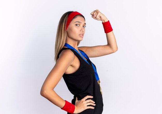 Giovane ragazza di forma fisica in abiti sportivi neri e fascia rossa con medaglia d'oro intorno al collo alzando il pugno guardando fiducioso in piedi sopra il muro bianco