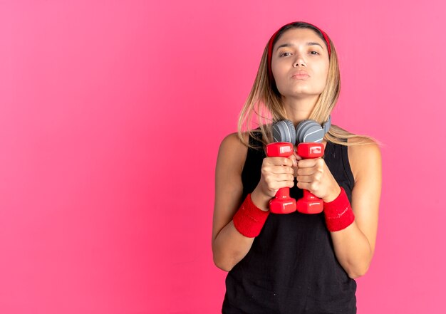 Giovane ragazza di forma fisica in abiti sportivi neri e fascia rossa facendo esercizi con manubri guardando fiduciosi sul rosa