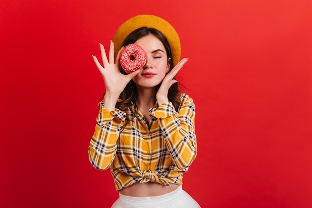 Giovane ragazza di ottimo umore copre gli occhi con ciambella alla fragola. Donna con labbra rosse in berretto in posa con gli occhi chiusi sulla parete isolata.