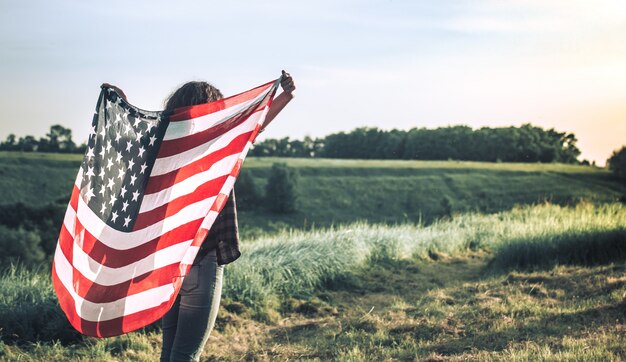 Giovane ragazza felice che corre e che salta spensierata con a braccia aperte sopra il campo di frumento. Tenendo la bandiera degli Stati Uniti.