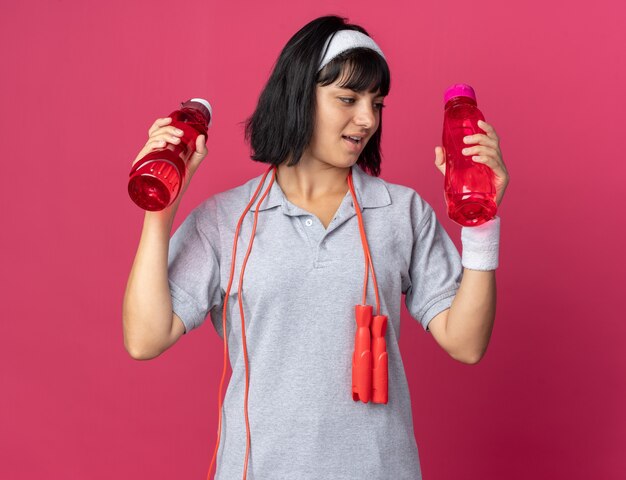 Giovane ragazza fitness che indossa fascia con corda per saltare intorno al collo tenendo due bottiglie d'acqua che sembrano confuse avendo dubbi