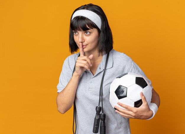 Giovane ragazza fitness che indossa fascia con corda per saltare intorno al collo tenendo in mano un pallone da calcio facendo gesto di silenzio con il dito sulle labbra in piedi su sfondo arancione