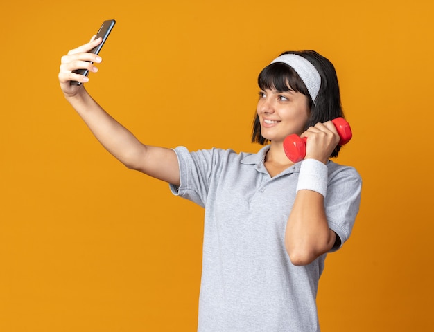 Giovane ragazza fitness che indossa la fascia che tiene il manubrio facendo selfie utilizzando lo smartphone sorridendo allegramente in piedi sopra l'arancia